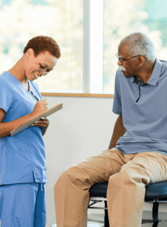 A physiotherapist in scrubs is talking to an elderly man sitting on an examination table and taking notes. 